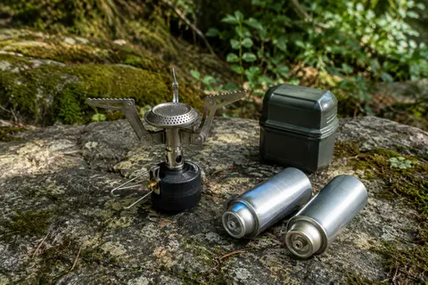 Réchaud de camping à quatre supports posé dans un sous-bois, avec deux cartouches perçables à côté.