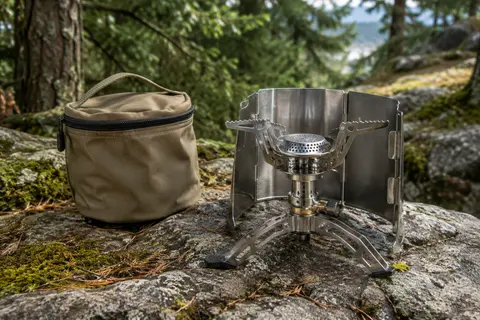 Réchaud de camping en acier avec stabilisateur et pare-vent, posé dans un sous-bois de montagne.