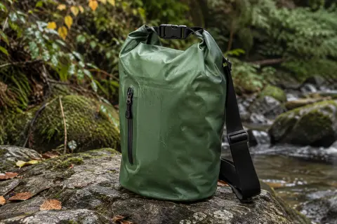 Sac étanche vert de 10 litres avec poche avant zippée et bandoulière, posé près d’un sous-bois.