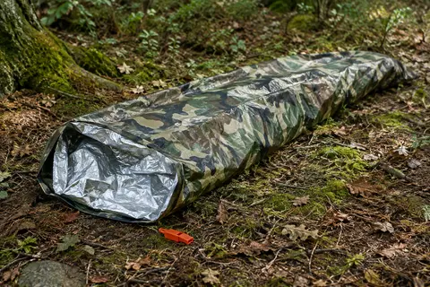 Sac de bivouac de survie camouflage déplié dans un sous-bois, avec son sifflet posé à côté