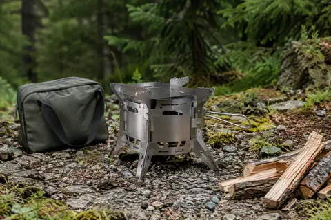 Réchaud de camping pliable LIXADA en acier inoxydable avec son sac, posé près de morceaux de bois en forêt.