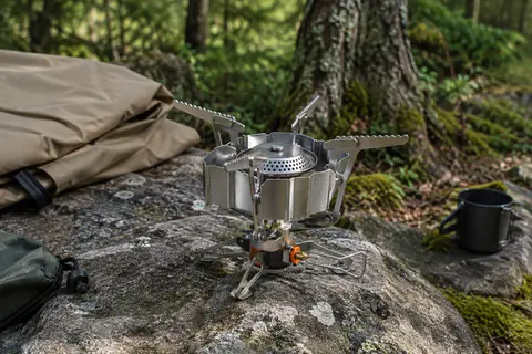 Réchaud de camping pliable en métal avec supports déployés, posé sur une pierre en sous-bois