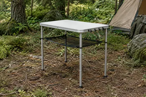 Table de camping blanche Sportneer avec pieds en aluminium, étagère en filet et crochets dans un sous-bois.