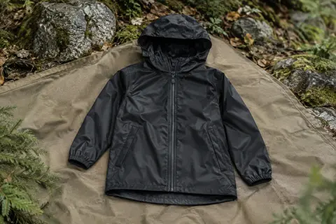 Veste de pluie noire pour enfant avec capuche, fermeture éclair et deux poches, posée dans un sous-bois.