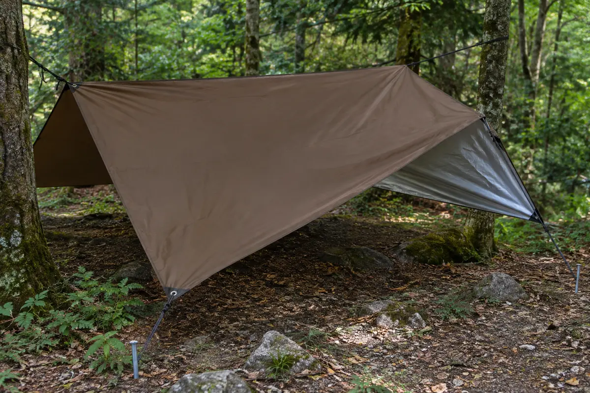 Bâche de bivouac marron tendue dans un sous-bois, avec ses cordes, ses piquets et sa toile argentée