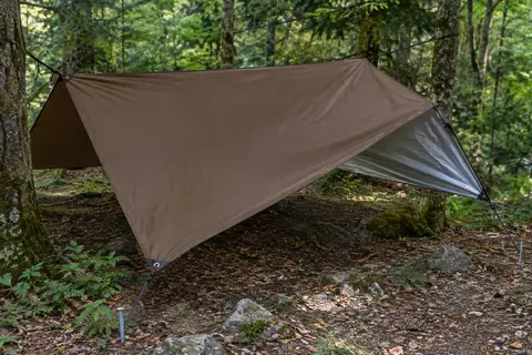 Bâche de bivouac marron tendue dans un sous-bois, avec ses cordes, ses piquets et sa toile argentée