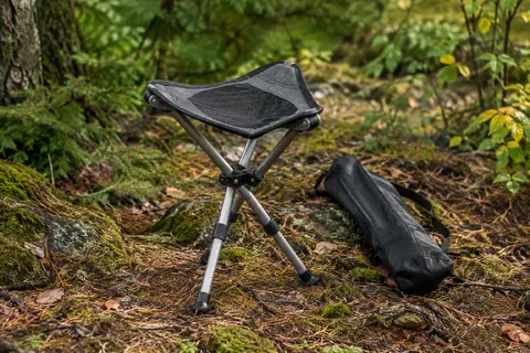 Tabouret pliant Walkstool noir et gris à trois pieds en aluminium, posé en sous-bois avec son sac.