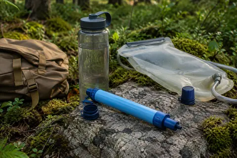 Paille filtrante portable bleue posée sur une pierre en sous-bois avec une bouteille et une poche à eau