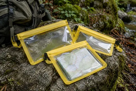 Trois pochettes étanches jaunes en PVC, dont une laisse voir une carte, posées sur une pierre en sous-bois