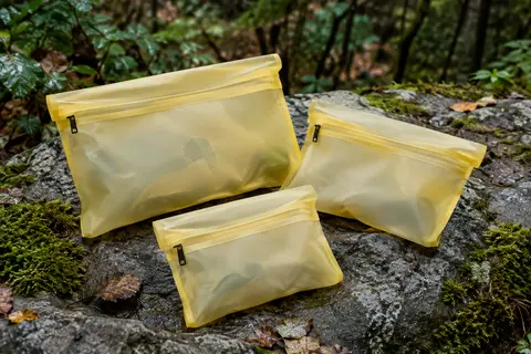 Trois pochettes étanches YUMQUA en PVC givré jaune, posées sur une pierre près d’un sous-bois.